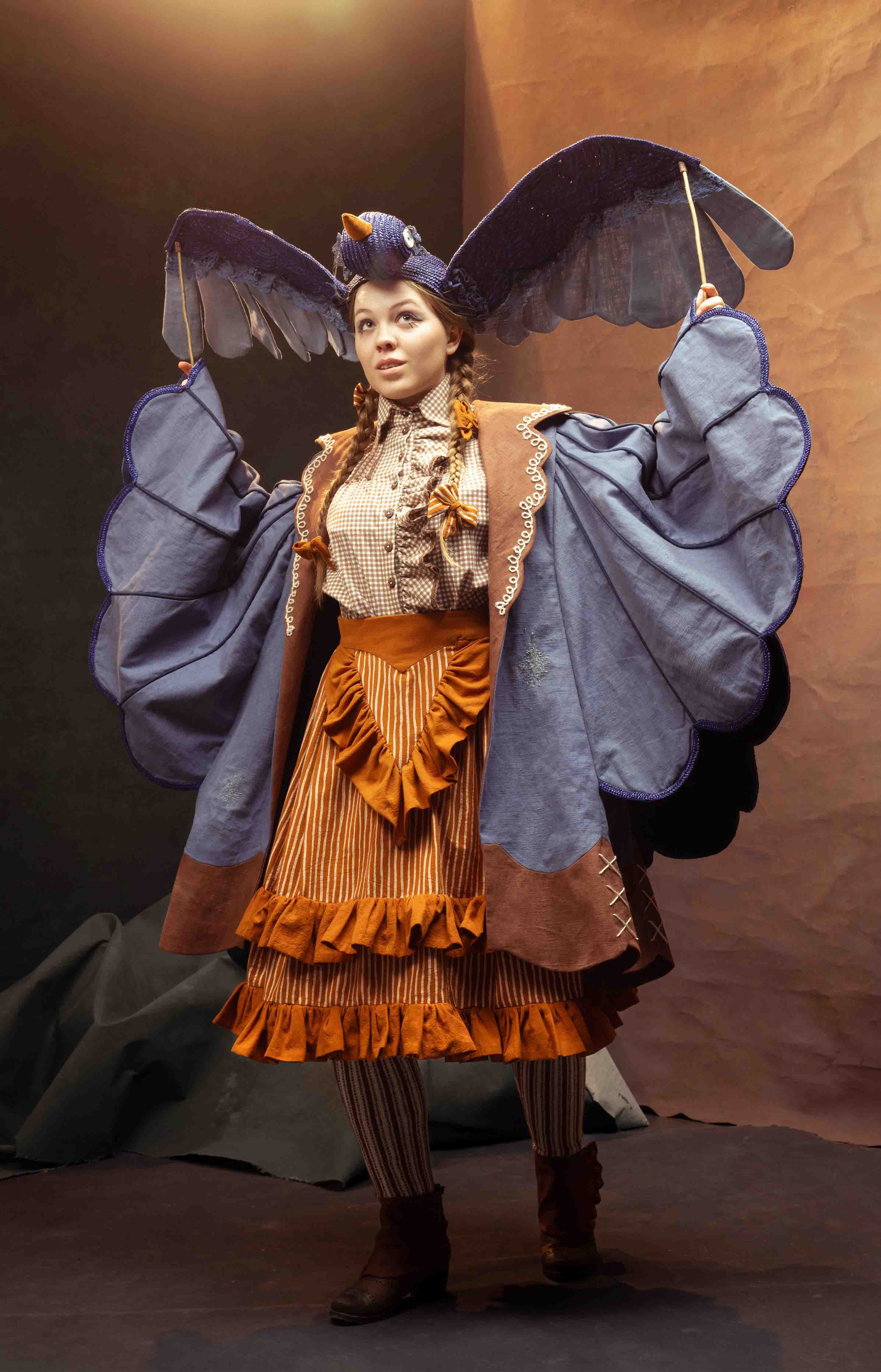 Costume for 'Beatrice' from Over The Garden Wall. A girl wears a bluebird puppet hat, a blue coat with large winged sleeves, an orange striped skirt and a brown gingham blouse.