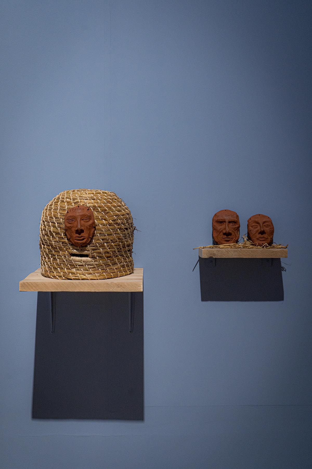 Two shelves. One displays a bee hive with a ceramic face attached, the other displays two ceramic faces.