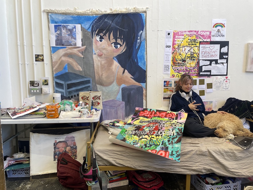 artist sat in their bed in the studio with their dog, they are surrounded by work-in-progress' artworks such as the painted rug and lisa simpson fingerpainting on the wall. the artist also has a large finger painting on paper covering their feet and the end of the bed. there is a desk with more art work on it infront of the wall tapestry and there is a JD vance image transfer showing below the desk 