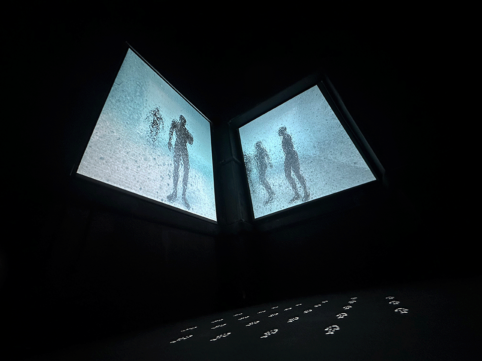 Low-angle view of two angled screens displaying hazy human figures above glowing floor projections.