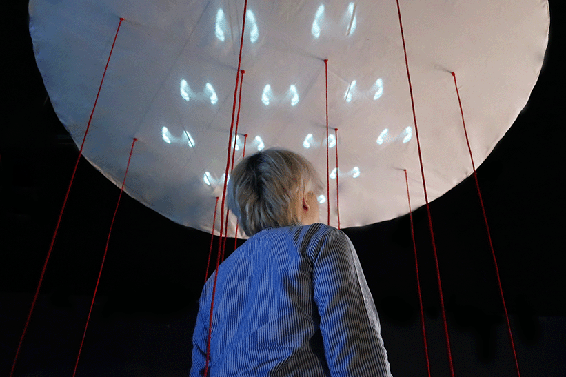 A participant gazes upward at a soft, white overhead structure where glowing footstep-like lights are embedded in a circular formation, with red threads connecting it to the floor.