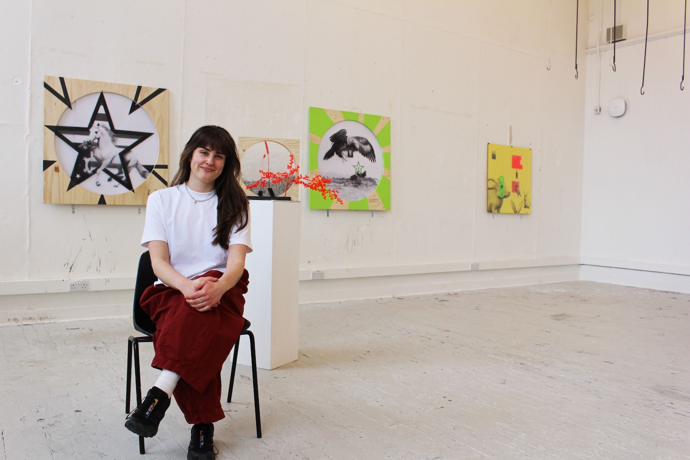 artist sat on a black chair, in a white room - in front of her works 