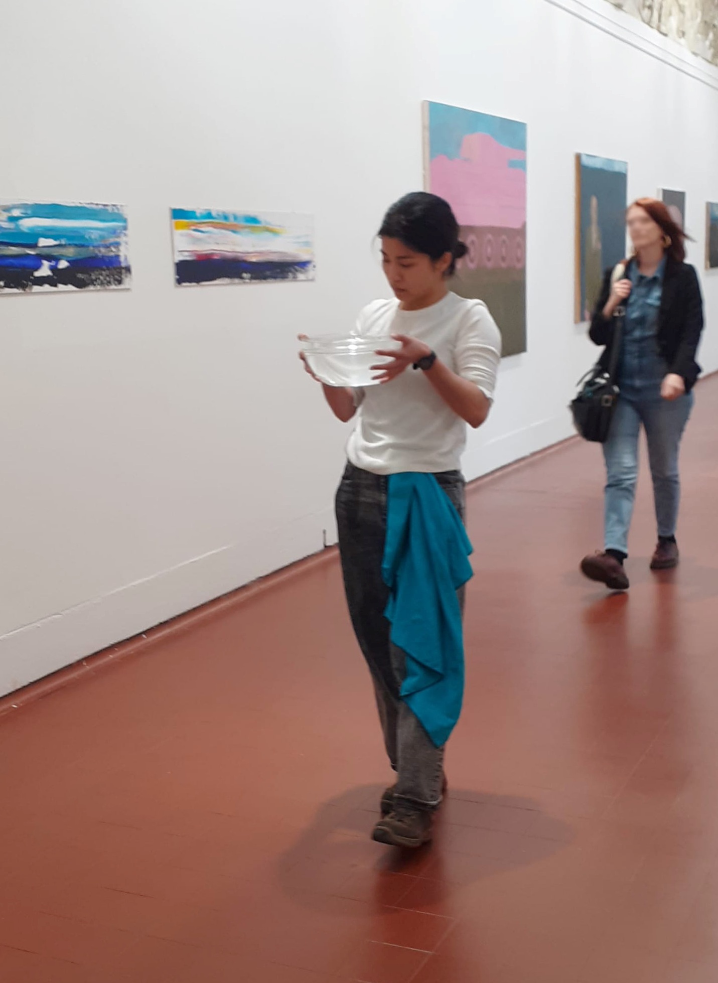 Lisa, the artist, walking down the corridor of Edinburgh College of Art's main building with a large glass bowl completely filled with water. Lisa is dressed in white long-sleeved shirt, dark jeans, hiking boots, watch, with a blue towel hanging from her waist. There are paintings on the white wall in her background and a visitor walking behind her. 