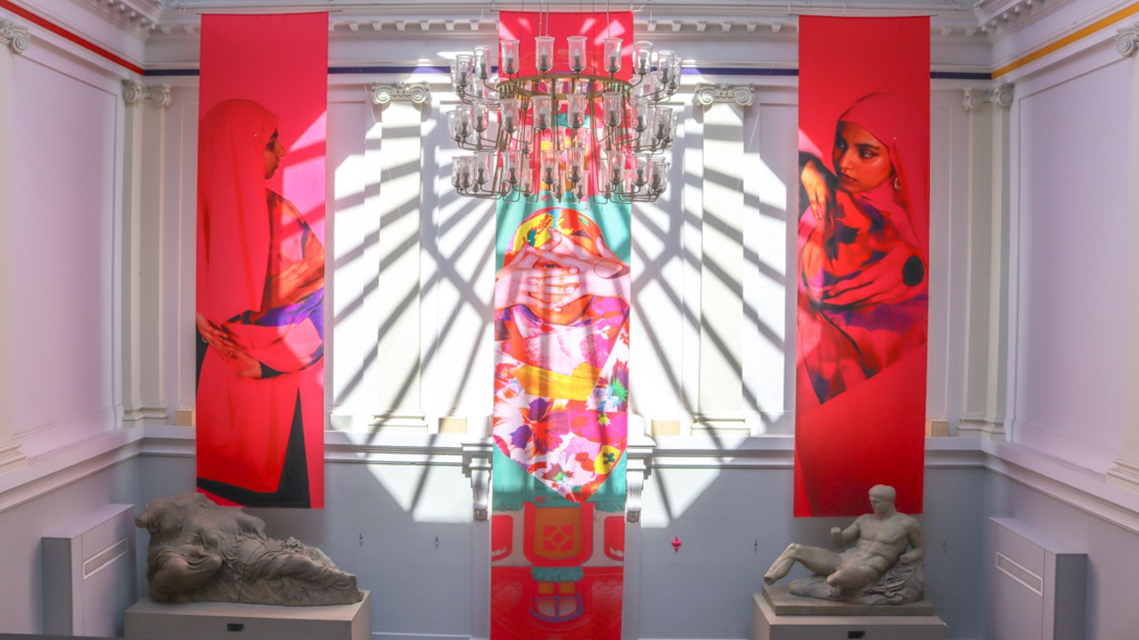 3 brightly coloured banners with 3 portrait images of the artist's face and shoulders (a Muslim woman wearing a floral scarf and pink scarves with images on them). Banners hang from the Edinburgh College of Art neoclassical stairwell which has a chandelier, columns and statue casts. The sunlight coming through the rough casts a magnificent shadow on the banners and the space