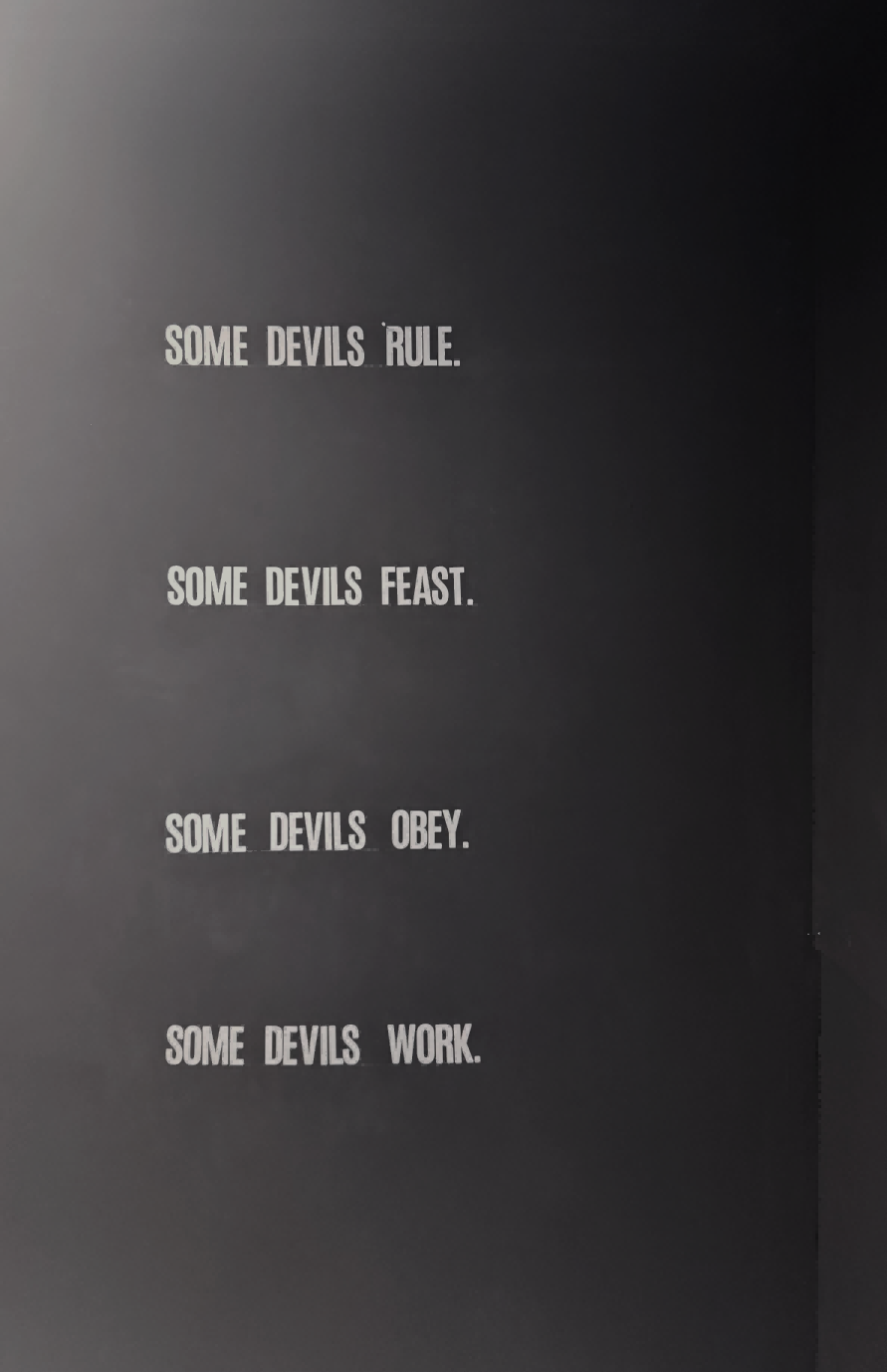 On the back wall, white vinyl text reads: “SOME DEVILS RULE. SOME DEVILS FEAST. SOME DEVILS OBEY. SOME DEVILS WORK.”