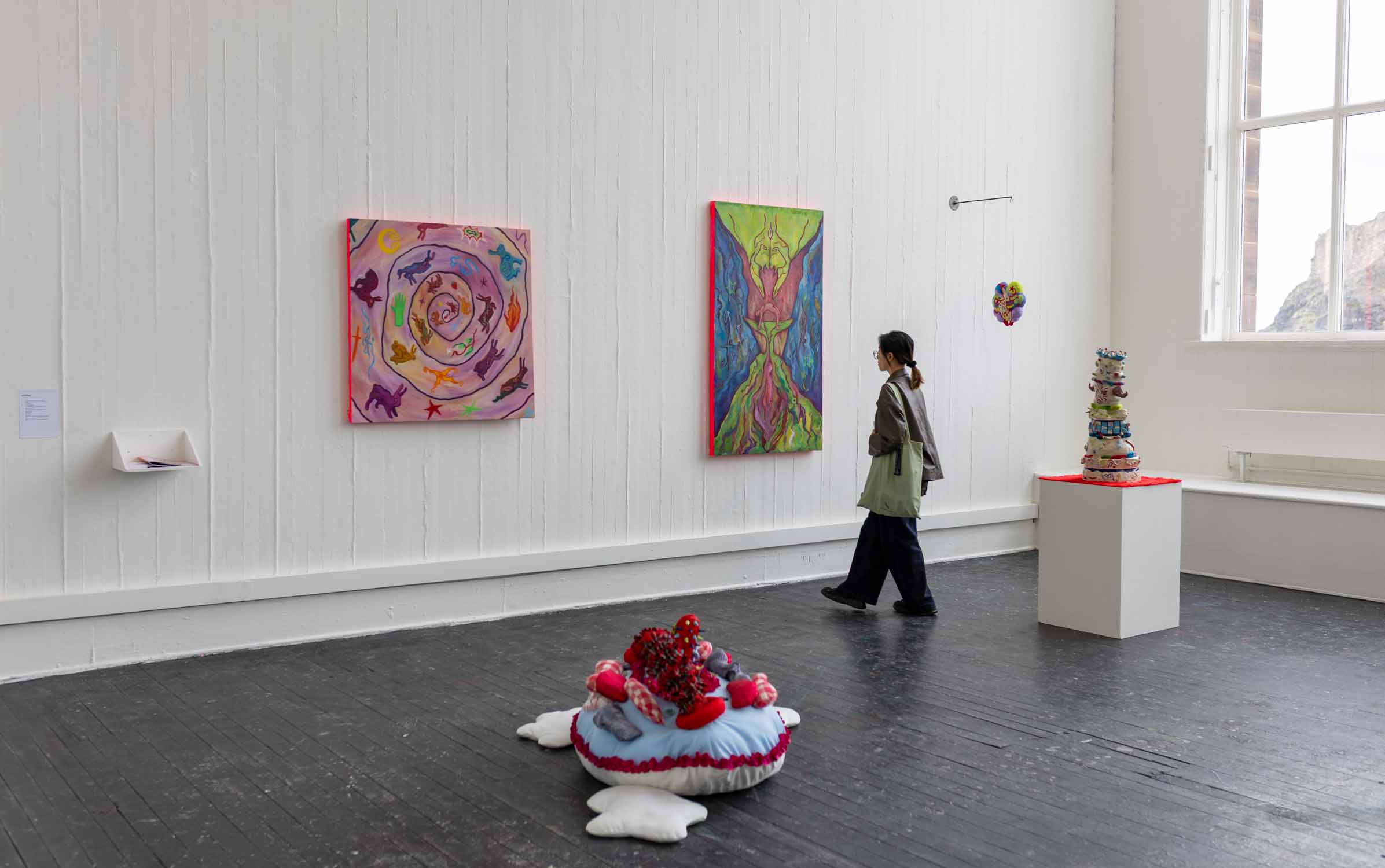 Degree Show Space with white walls and window. An audience is looking at the paintings on the wall, surrounded by colourful sculptures in the space. 