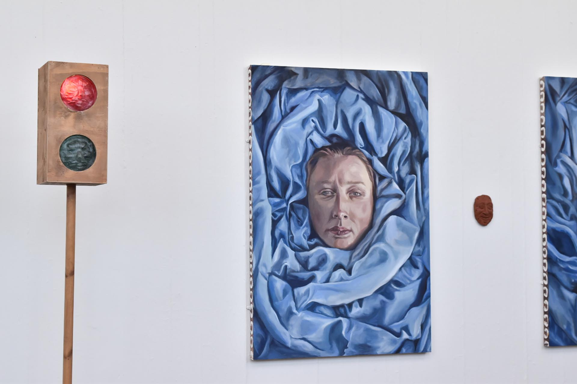 An image with two paintings mounted on a white wall containing a woman's portrait wrapped in blue fabric. between the paintings is a ceramic face. A traffic light is in the foreground of the image. 