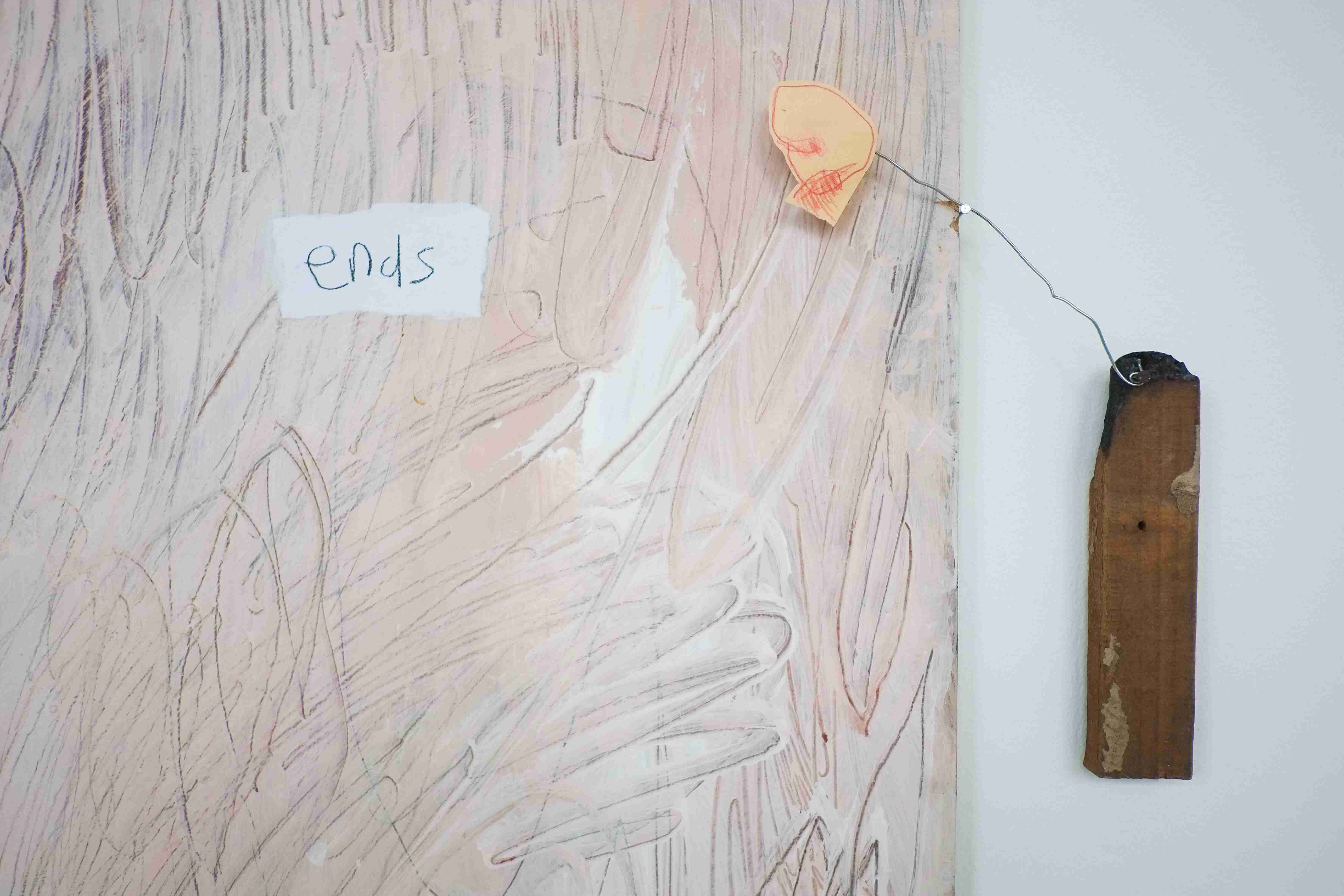 Close up of a light pink painting which reads 'ends'. It is accompanied by a sculptural figure made from wood, wire and paper to its side