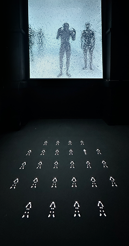 A wall-sized frosted screen displays large silhouettes above a floor grid of glowing human icons.