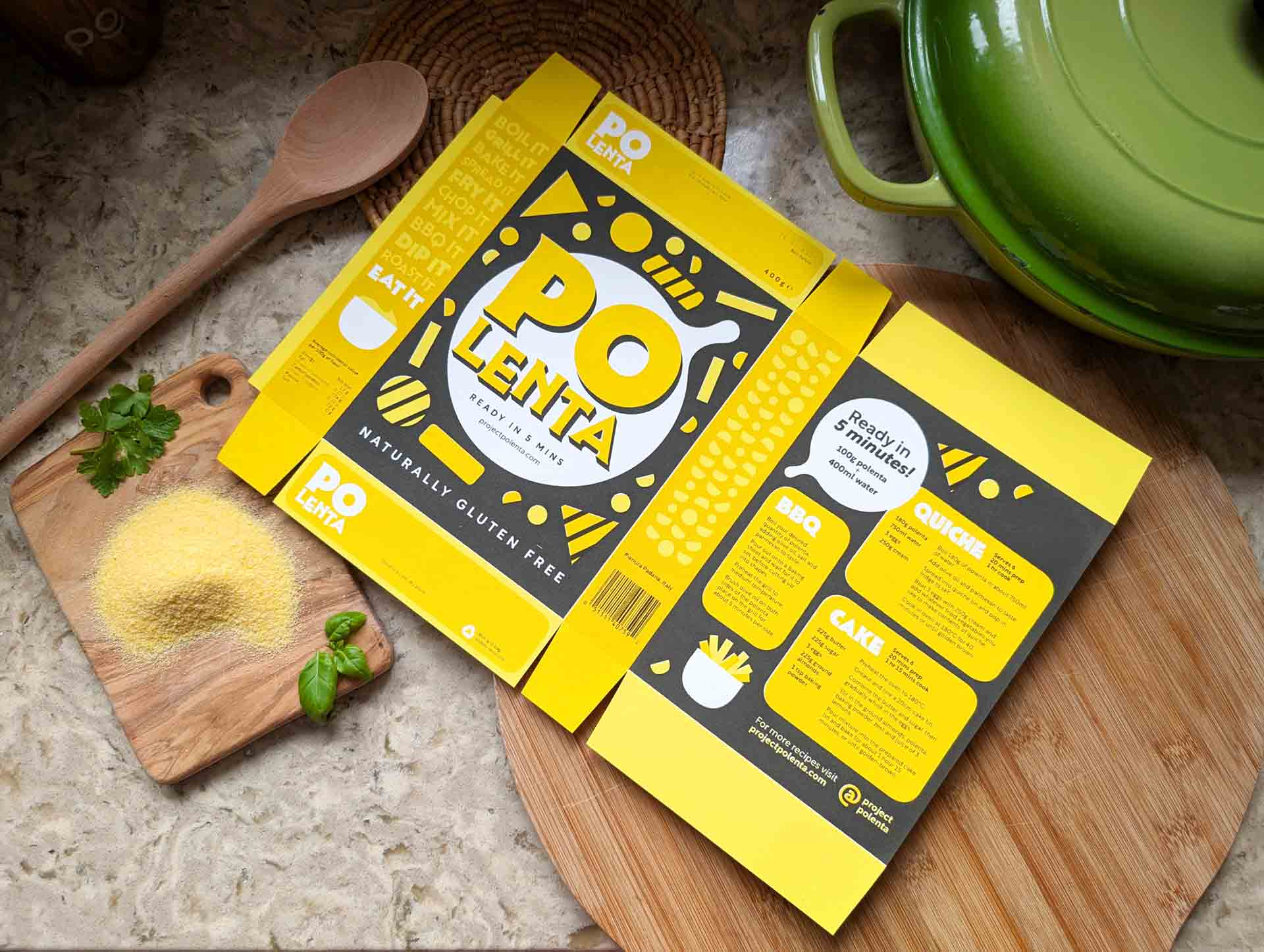 Unfolded polenta packaging on kitchen counter, surrounded by green crockery, a wooden spoon and chopping boards with basil leaves.