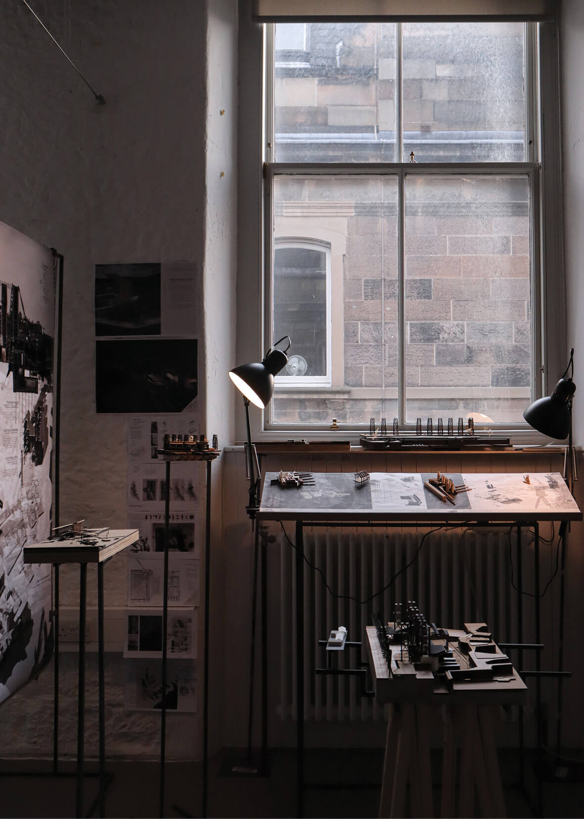 Photograph of an architecture studio workspace illuminated by soft natural light and two desk lamps. A drafting table sits beneath a tall sash window, displaying conceptual drawings and small-scale models. The surrounding walls are pinned with sketches and reference images, while additional presentation models are arranged on slender stands, creating an intimate and focused design environment.