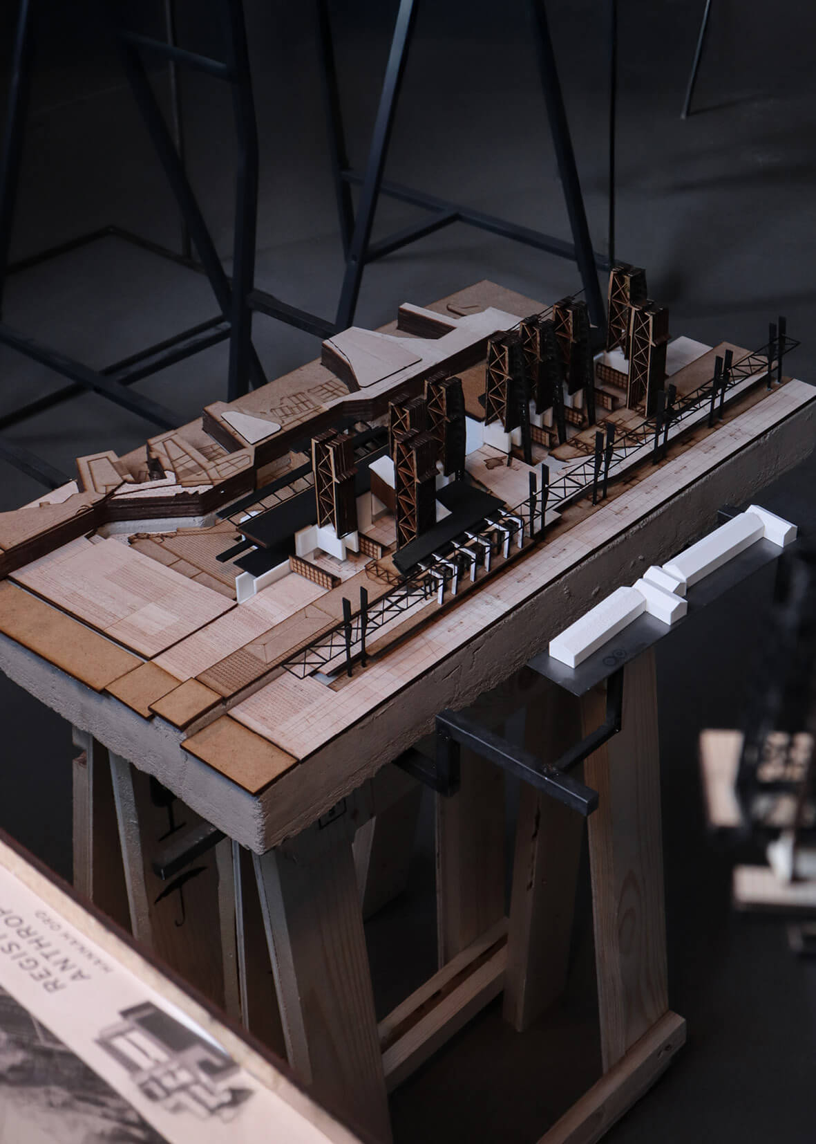 A detailed architectural model showcasing a linear industrial structure with vertical trusses and layered materials, displayed on a wooden stand. The model incorporates varied textures and tones in wood, card, and white elements to represent a complex site layout with infrastructure and built forms
