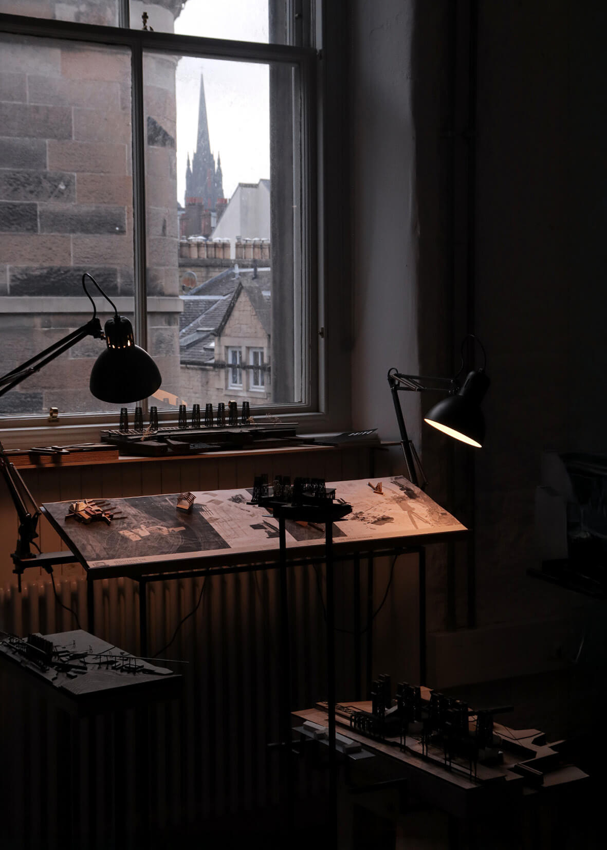 Moody interior photograph of an architecture studio with small-scale models and drawings illuminated by desk lamps. The scene captures a quiet workspace beneath a window overlooking historic Edinburgh rooftops, with the spire of the Hub visible in the distance. The lighting emphasizes the tactile quality of the models.