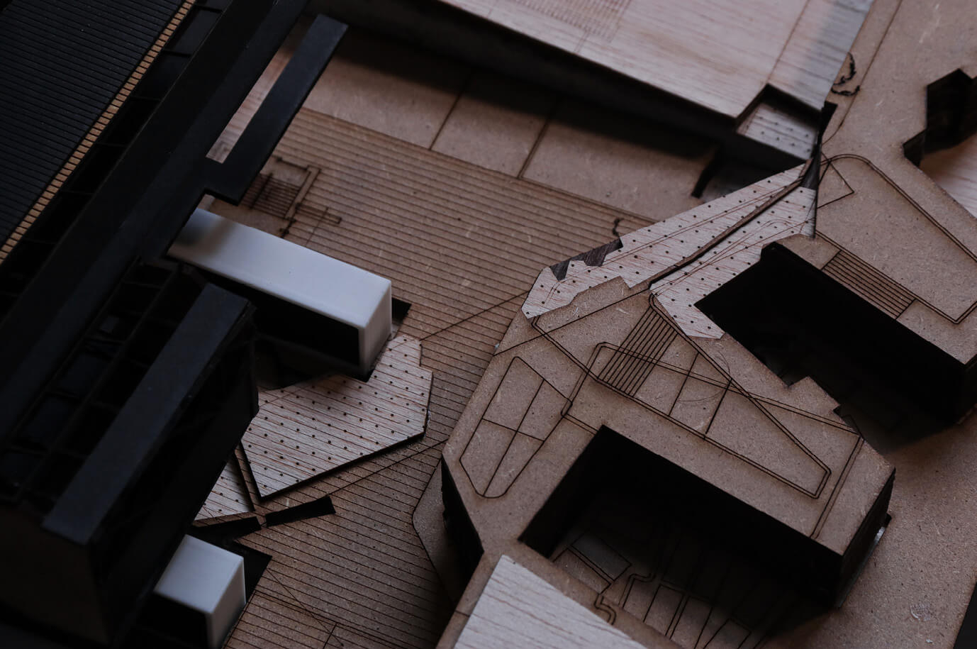 Close-up detail of a laser-cut architectural model made from layered wood, card, and acrylic. The image focuses on intricate engraved lines and cut-out building volumes, showing detailed topography, circulation paths, and user junctions.