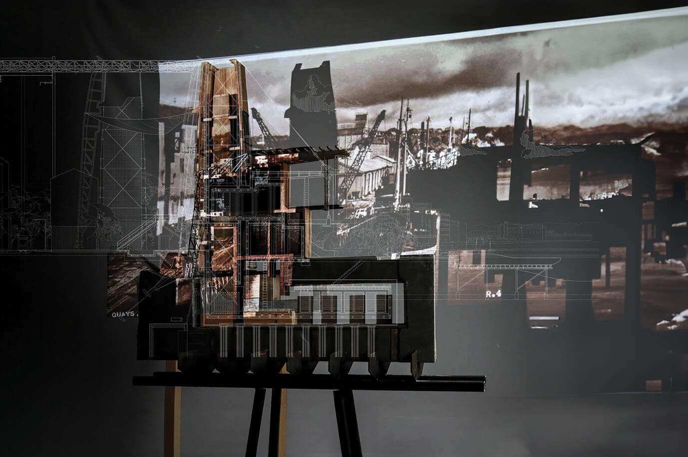 Photograph of a physical architectural section model on a stand, overlaid with a projected technical drawing and historic dockyard imagery. The model, detailed with internal partitions and structural systems, is lit dramatically against a dark backdrop. The projection merges hand-drawn section lines with sepia-toned visuals of cranes and shipyards, creating a layered composition that connects the design to its post-industrial context and infrastructural lineage.