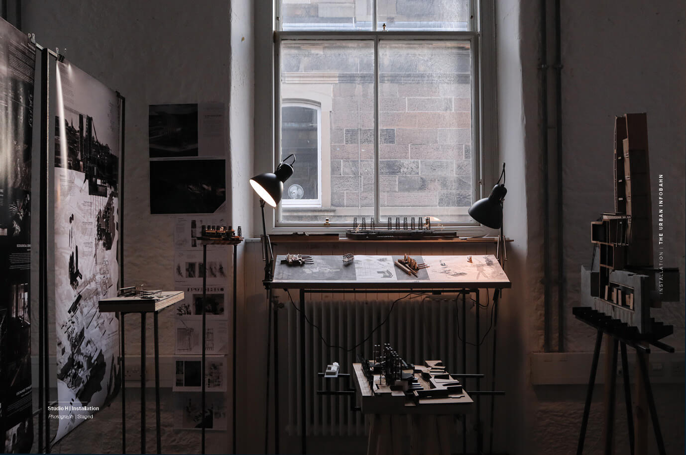 Photograph of a studio installation showing a workspace with architectural models, printed drawings, and presentation boards. A central desk sits beneath a large window, lit by two desk lamps. Models are arranged on tables and stands around the room, and large posters with project visuals are pinned to the wall on the left. The image documents the layout and materials used for the architectural presentation.