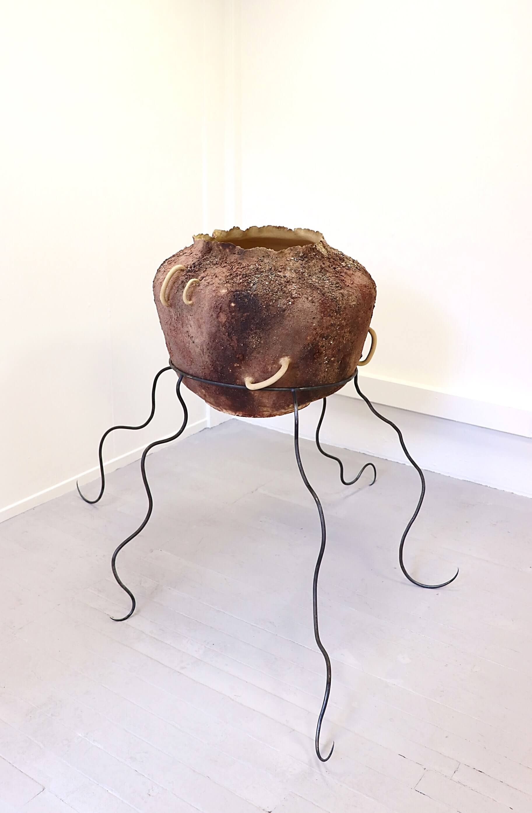 Large ceramic pot embedded with crushed rocks, with an unusual slanted handle and standing on 5 curved steel legs.