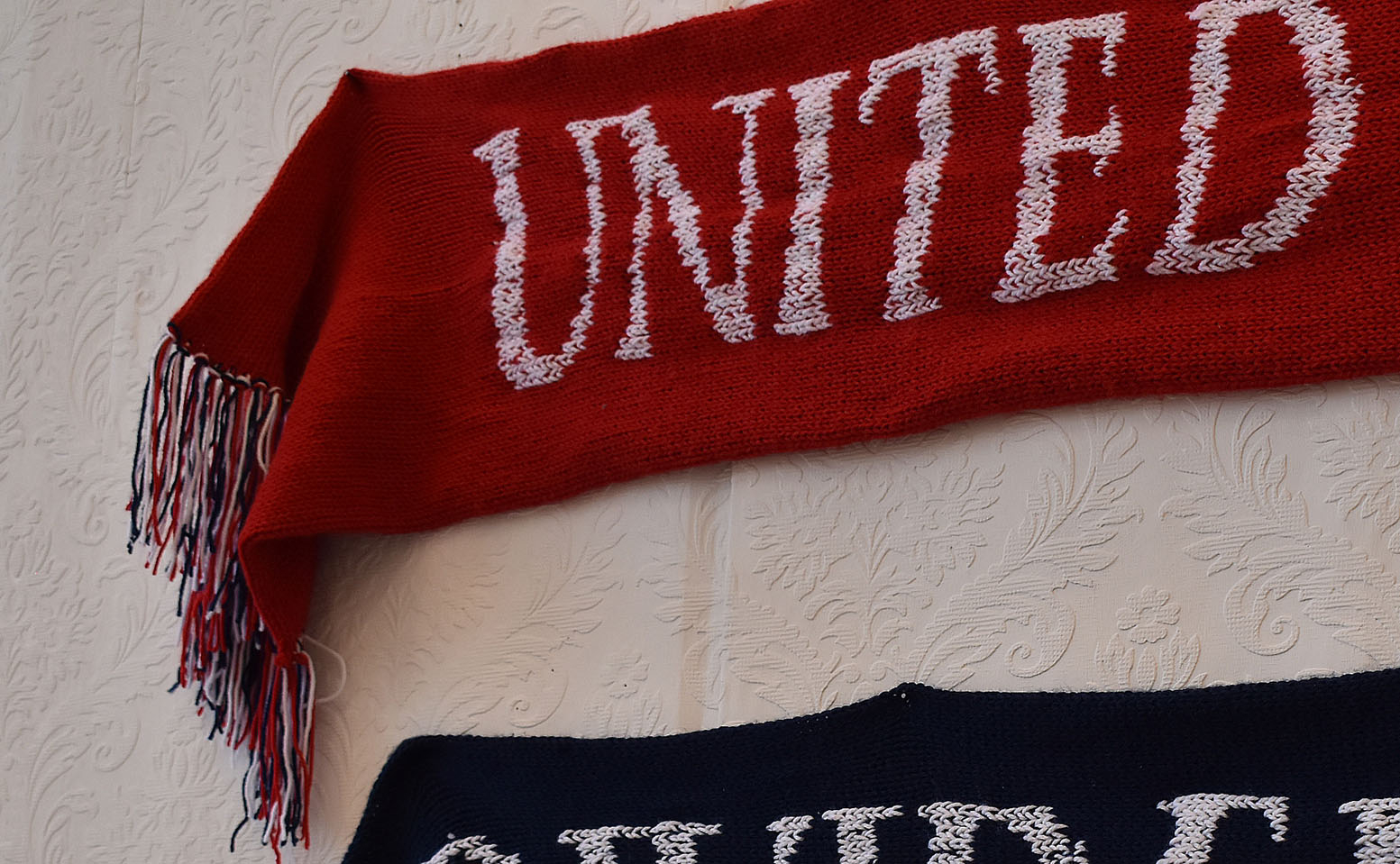 Red knitted scarf with text 'UNITED' stitched on. Scarf hung on wallpapered wall.