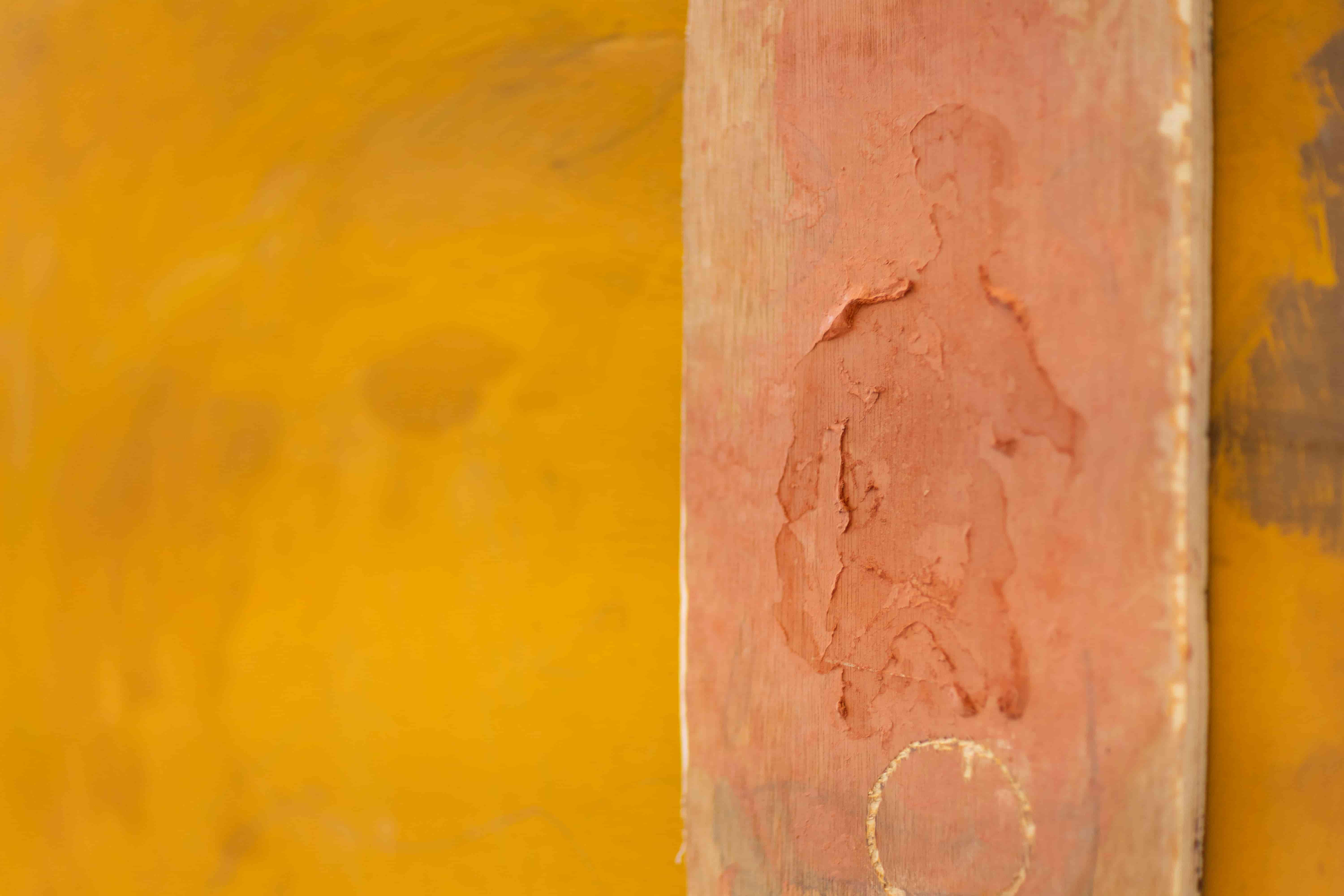 Close up of a vibrant golden-yellow painting on wood. The outline of a man in red clay can be seen on a wood offcut placed on top of it.