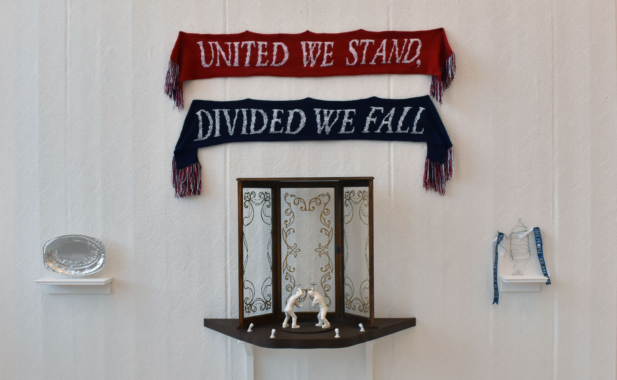 wall of 3 sculptural works arranged as a triptych, two scarves, red and blue are above the central work