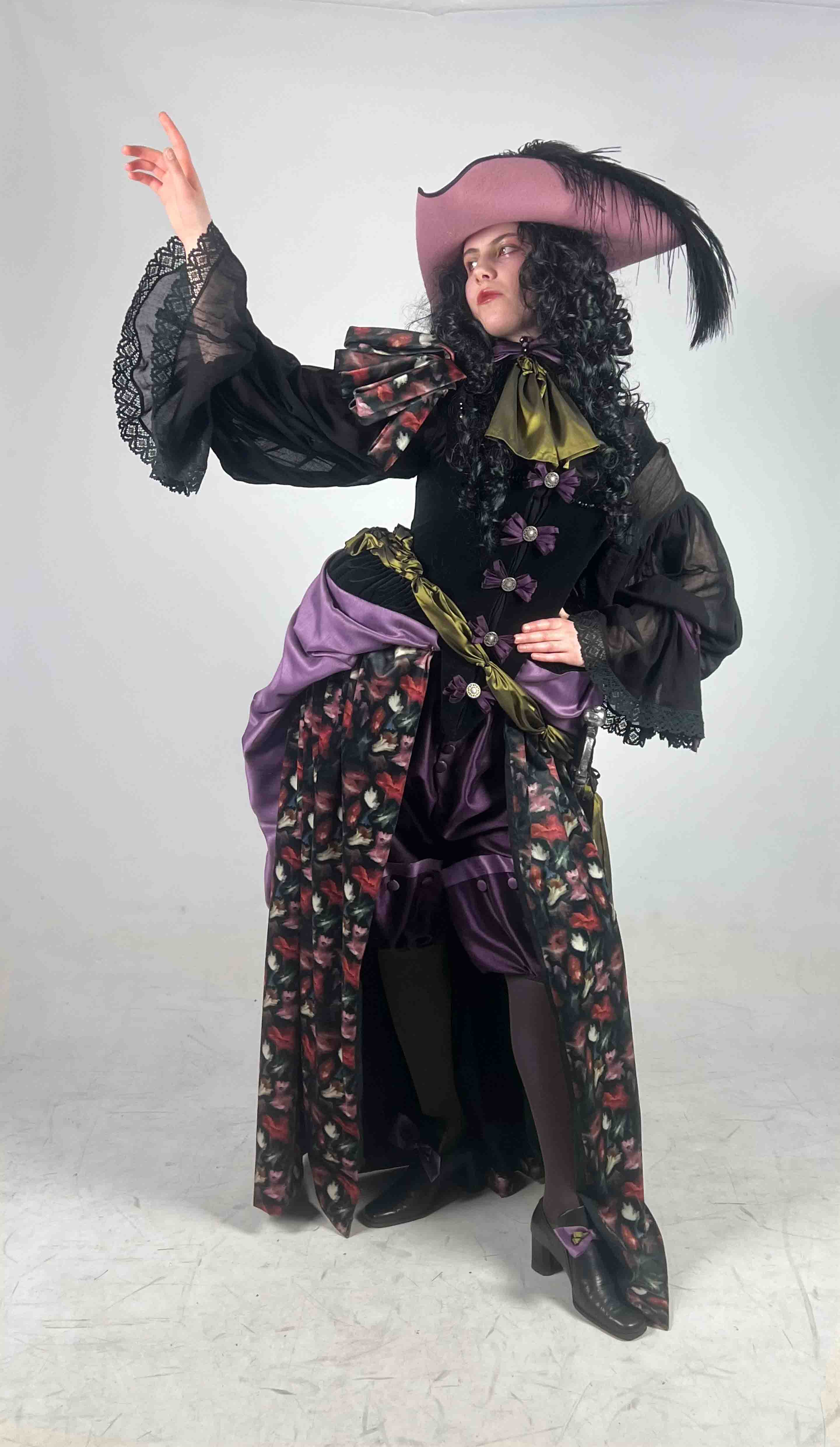 A woman dressed in a pink tricorne, black curly wig, black bodice and shirt, and floral velvet skirt.