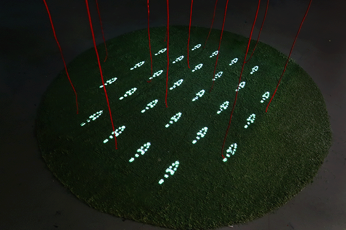 Top-down view of a circular patch of artificial grass displaying a precise grid of glowing digital footprints, intersected by red hanging strings.