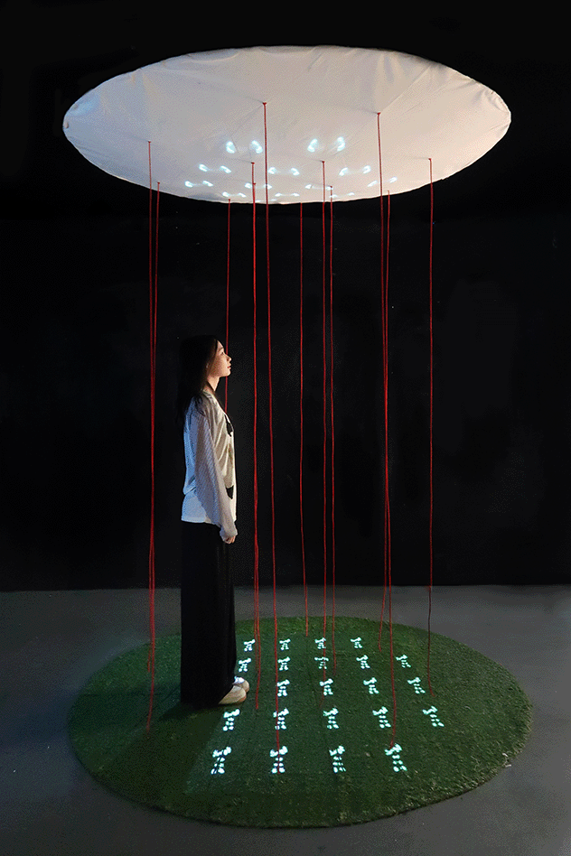 An installation featuring a large white circular canopy suspended above, from which red strings hang vertically around a standing person; illuminated footprints are projected in a grid on the circular green turf below.