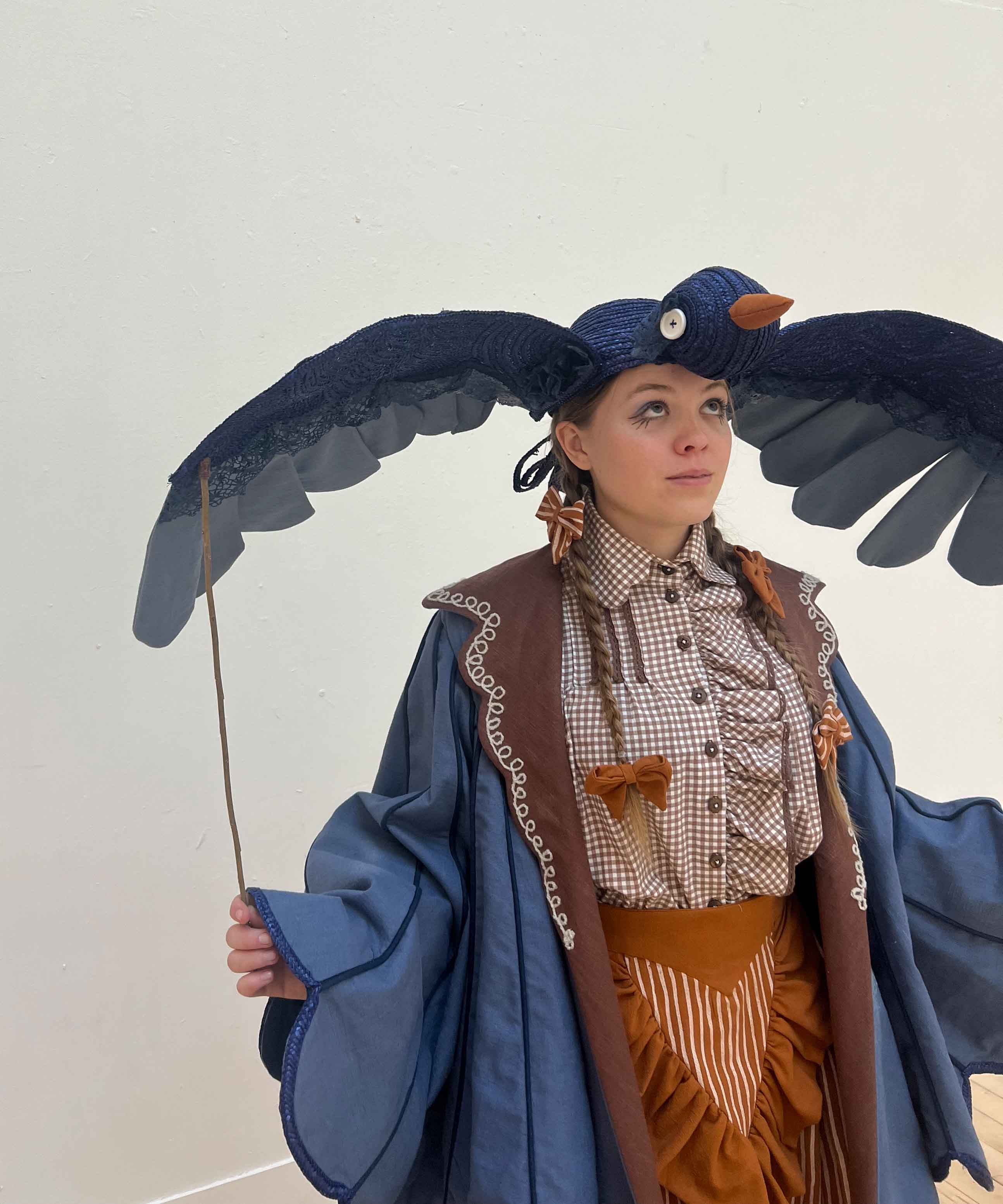 Costume for 'Beatrice' - a girl wears a bluebird puppet hat made of straw and linen, a large blue coat with brown lapels, a brown gingham shirt and striped orange skirt.