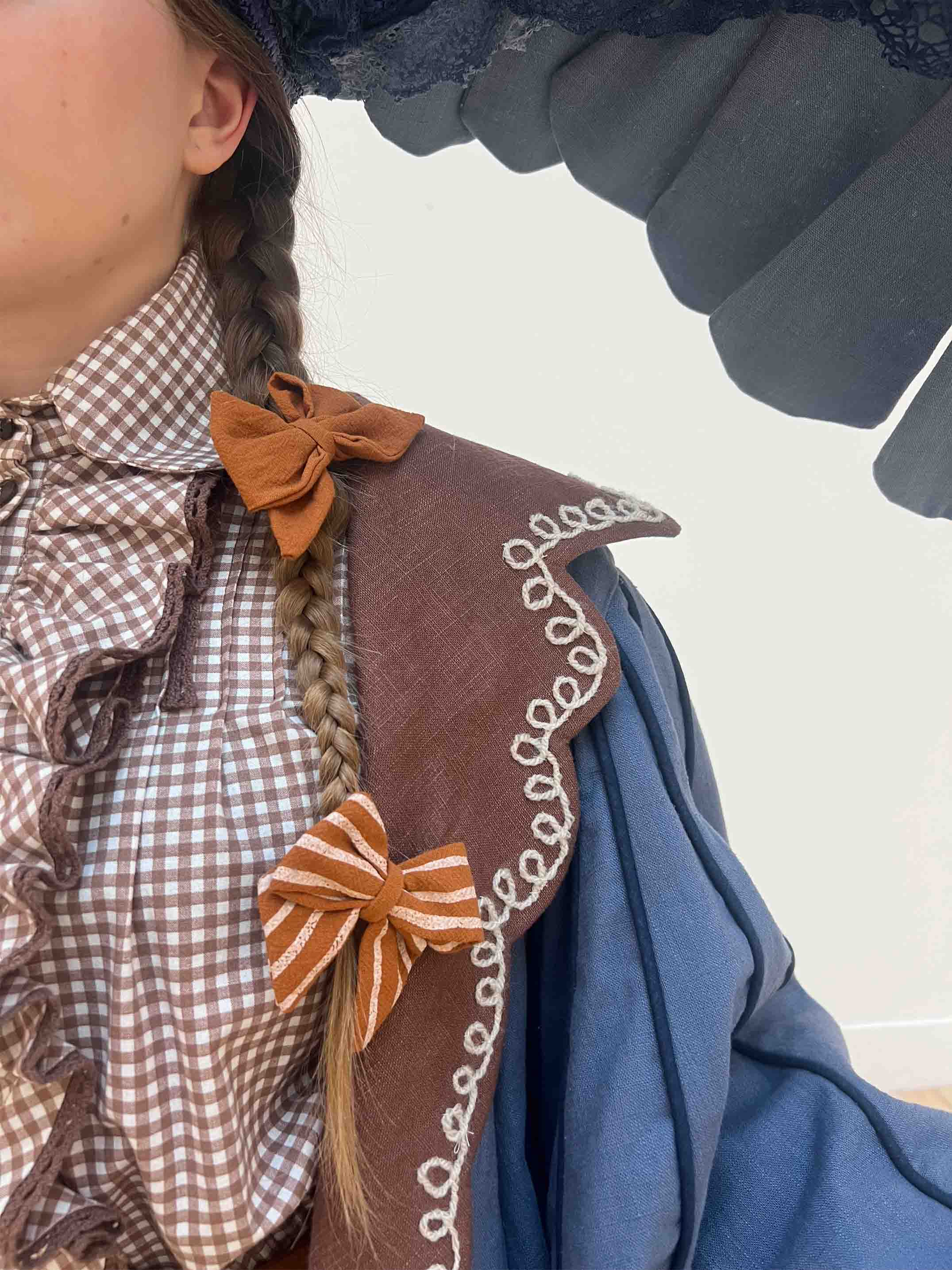Costume for Beatrice - zoomed in detail shot of the white yarn embellishment on the brown lapel of the coat. Also visible are the orange bows in the braids of the models hair.