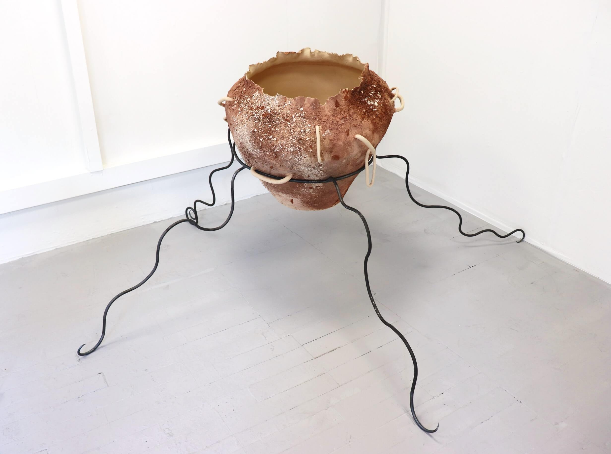 An inverted traingle shaped ceramic pot embedded with rocks, elevated by bendy steel stands which appear like creature legs.
