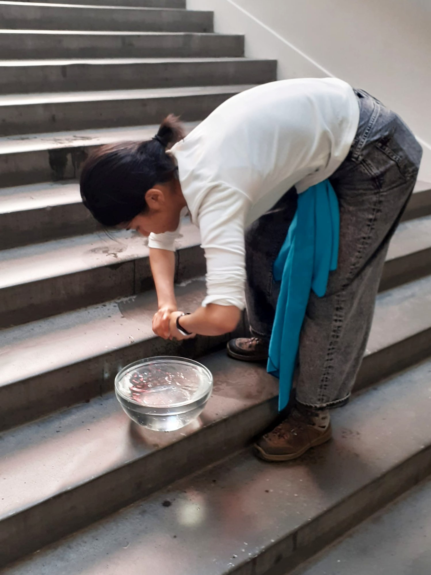 Lisa, the artist, bending down and looking down to the glass bowl filled with water, placed on the stairs. She is draining as much water as possible from a white sponge with both hands, twisting it tightly onto the glass bowl, adding the spilled water back into the bowl. 