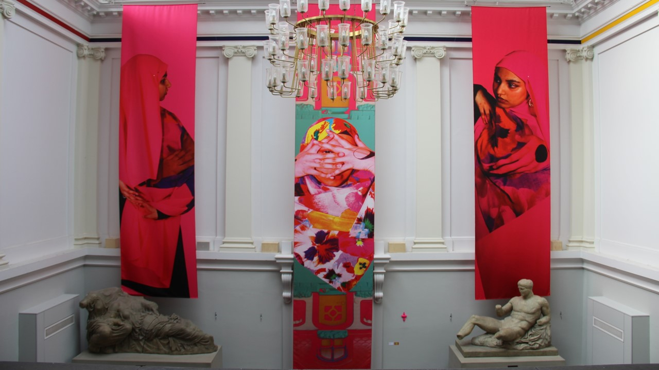 3 brightly coloured banners with 3 portrait images of the artist's face and shoulders (a Muslim woman wearing a floral scarf and pink scarves with images on them). Banners hang from the Edinburgh College of Art neoclassical stairwell which has a chandelier, columns and statue casts.