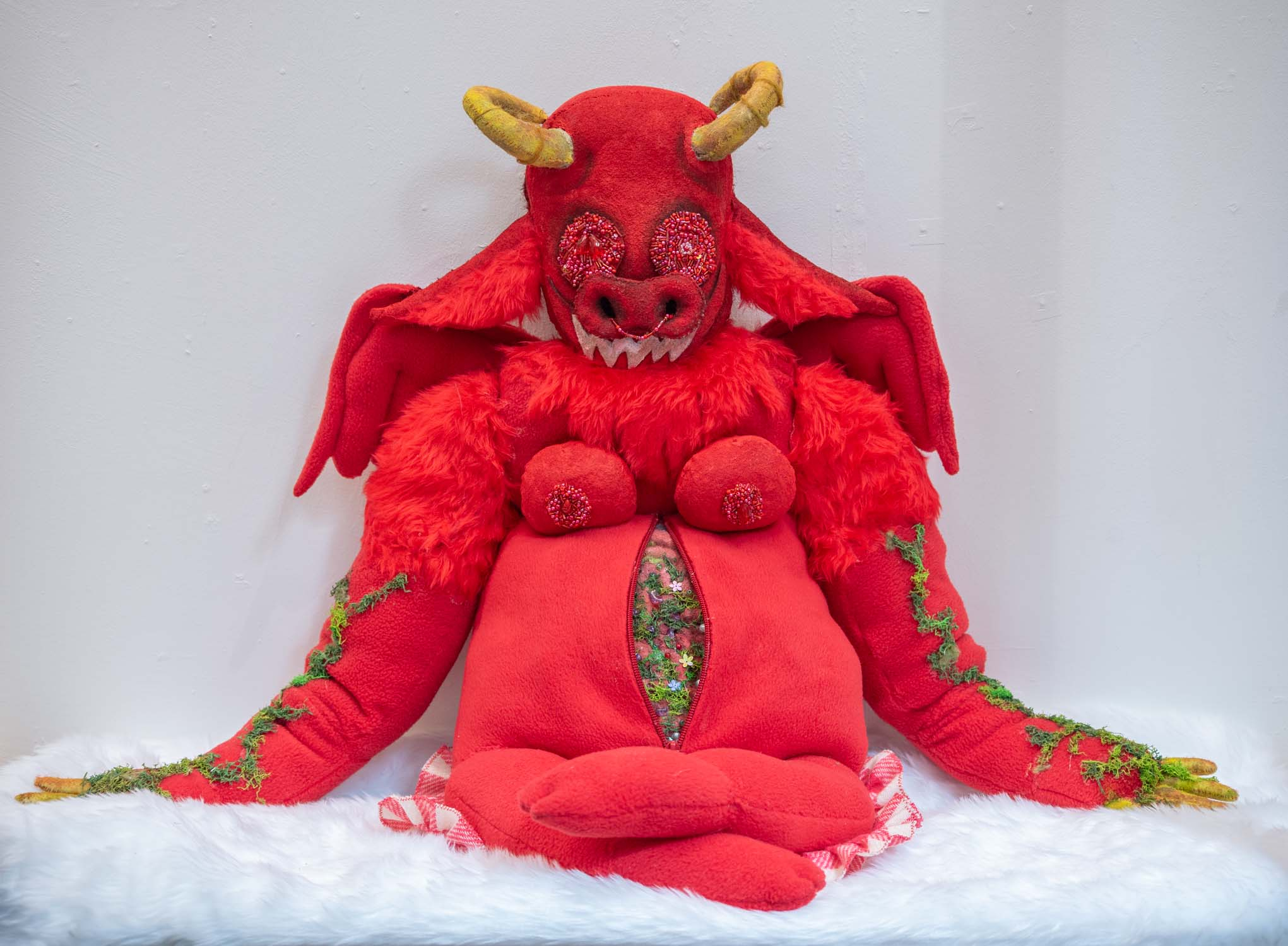 Soft sculpture of a red horned creature with bedazzled eyes and nipples. There is a zipper on its belly that opens to reveal artificial lichen. The creature is grinning in a cross-legged pose. 