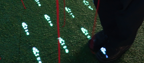 Close-up view of a person's feet standing among glowing footprint projections on green turf, partially obscured by vertical red strings.