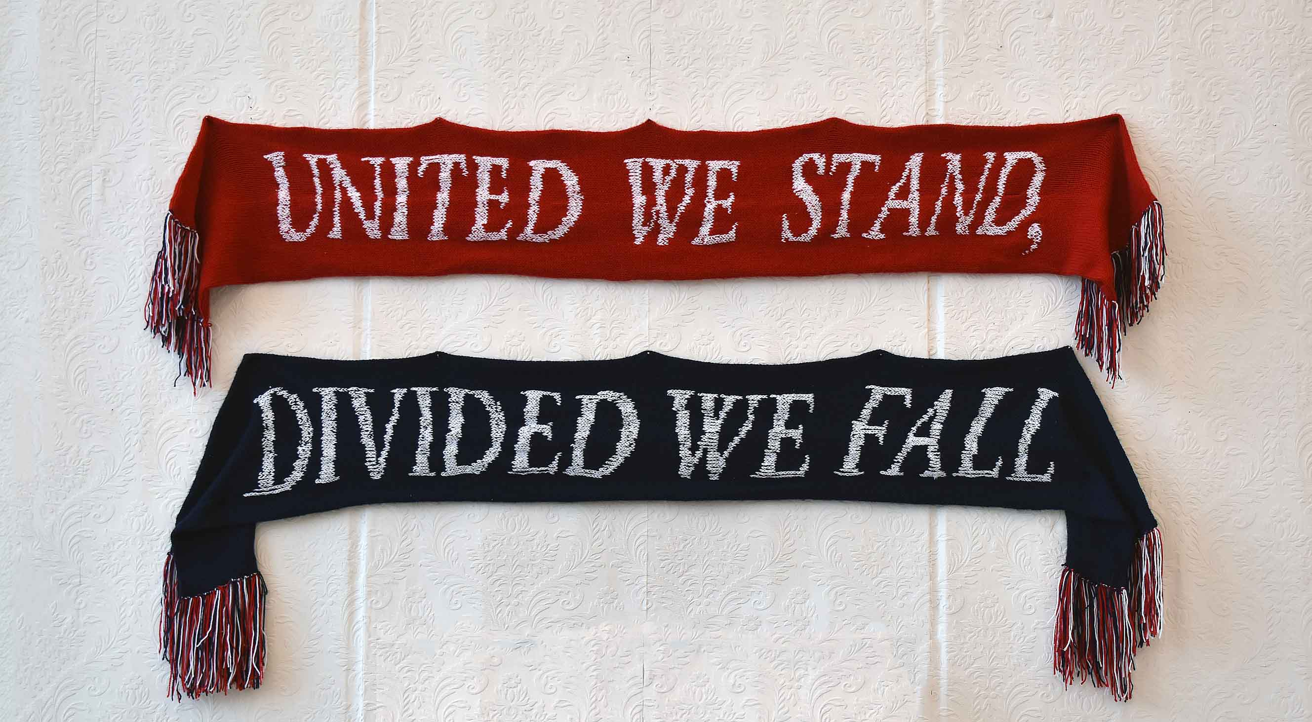 Two knitted scarves, one red reading 'UNITED WE STAND' with red, white and blue tassels, and below one navy blue reading 'DIVIDED WE FALL' with the same tassels