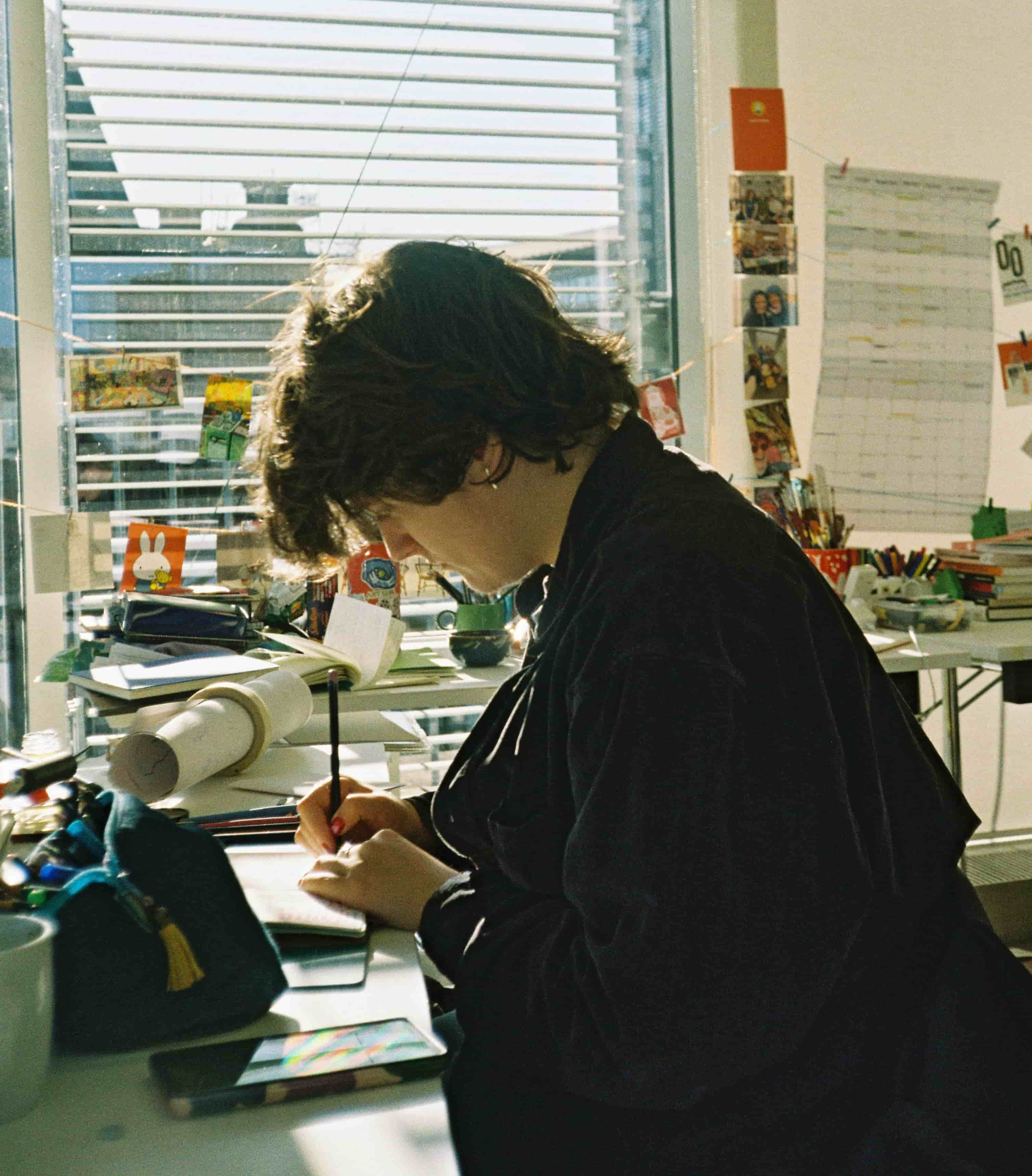 Mollie sits at a messy desk in a brightly lit studio drawing