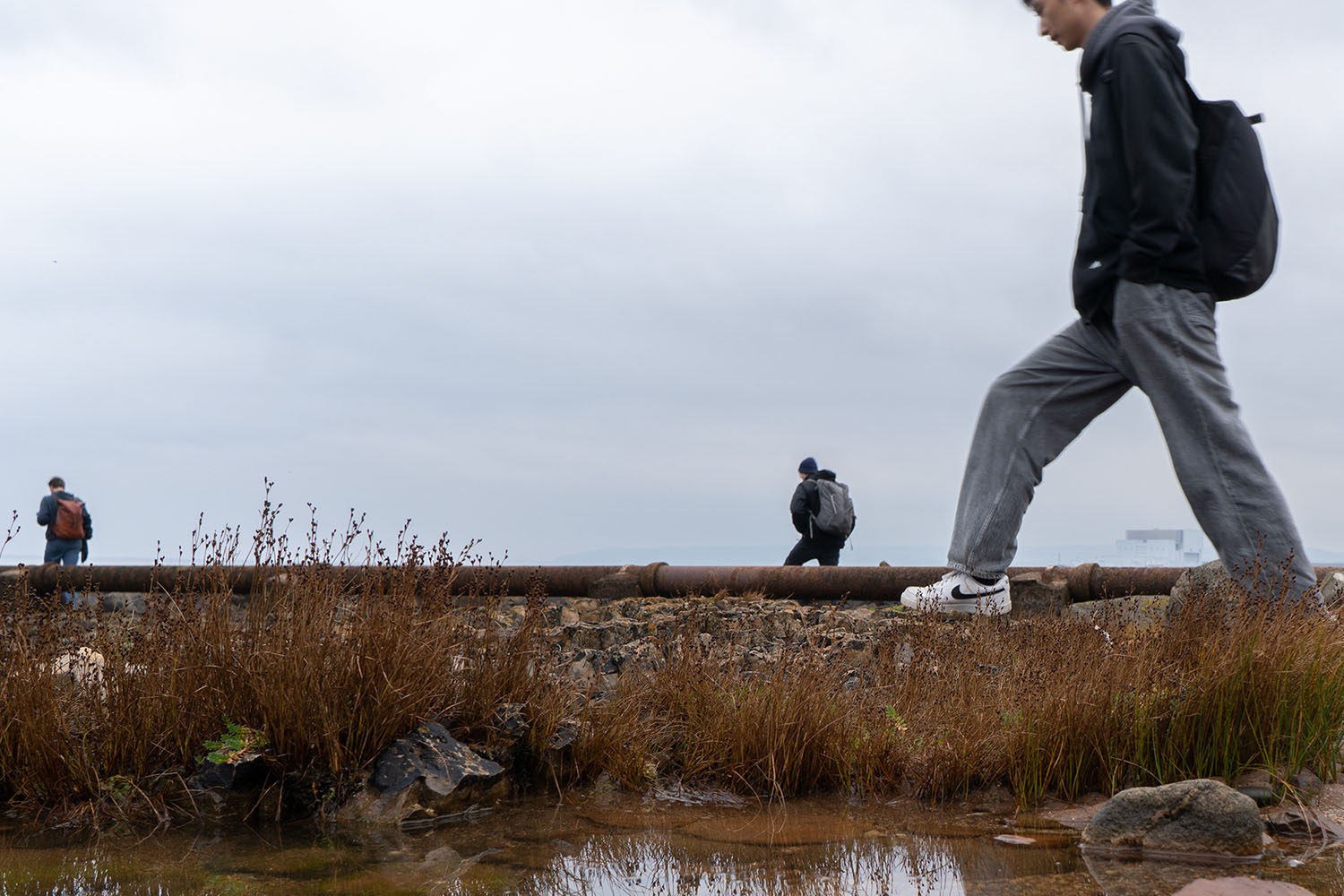 A student is walking along the edge of a shallow waterway. Two people are far in the distance and looking away from the camera.
